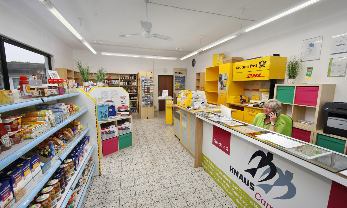 At KNAUS Campingpark Wingst, a cashier converses on the phone in a small campsite shop, surrounded by shelves of groceries and a postal service counter.