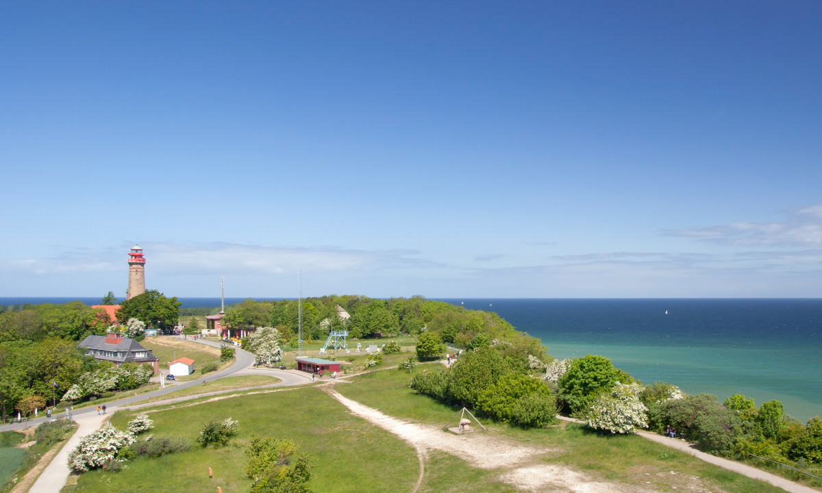 At KNAUS Campingpark Rügen, a lighthouse stands amidst lush vegetation and blossoms, adjacent to a coastal trail with a picturesque ocean view.