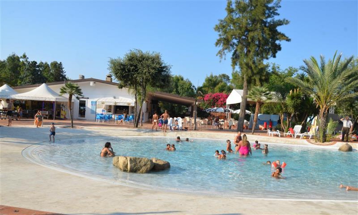 At Ultima Spiaggia, individuals are swimming and unwinding in a spacious outdoor pool encircled by trees and structures.