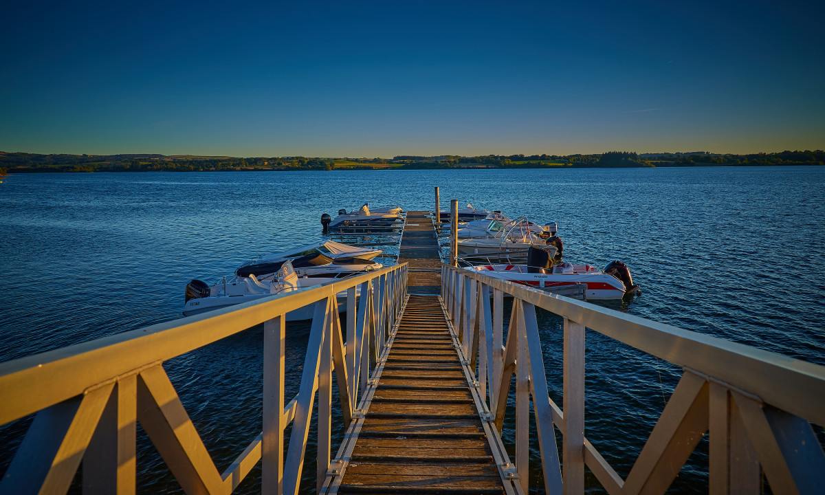 At Terrasses du Lac, a wooden dock extends towards a number of small vessels moored on a serene lake at sunset, with distant hills visible in the background.