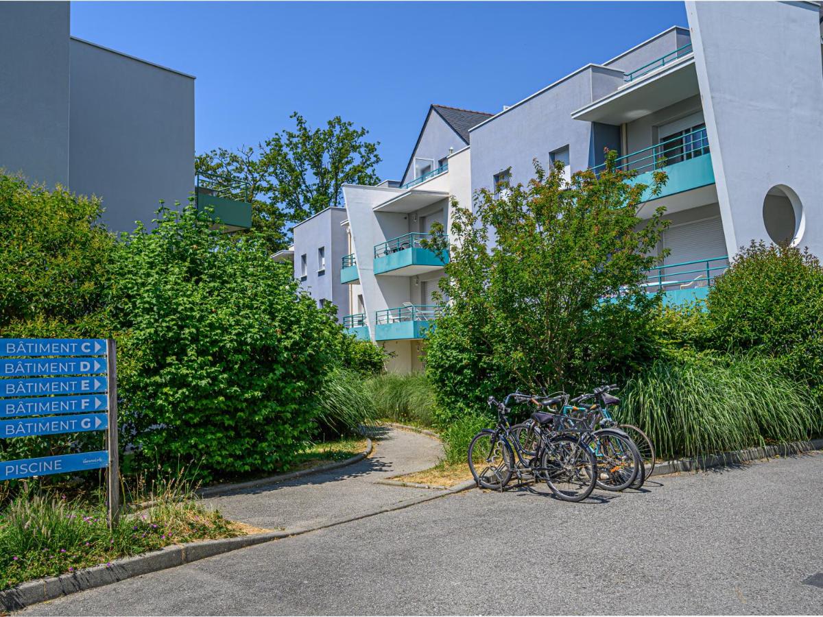 Bicycles are parked near a pathway, surrounded by greenery, with modern buildings in the background at Ker Goh Lenn
