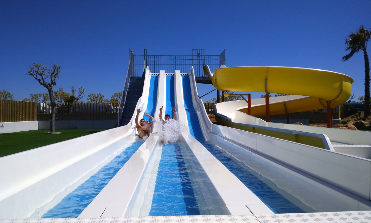 People sliding down water slides, raising their hands, in a pool area with clear skies at Resort Els Pins